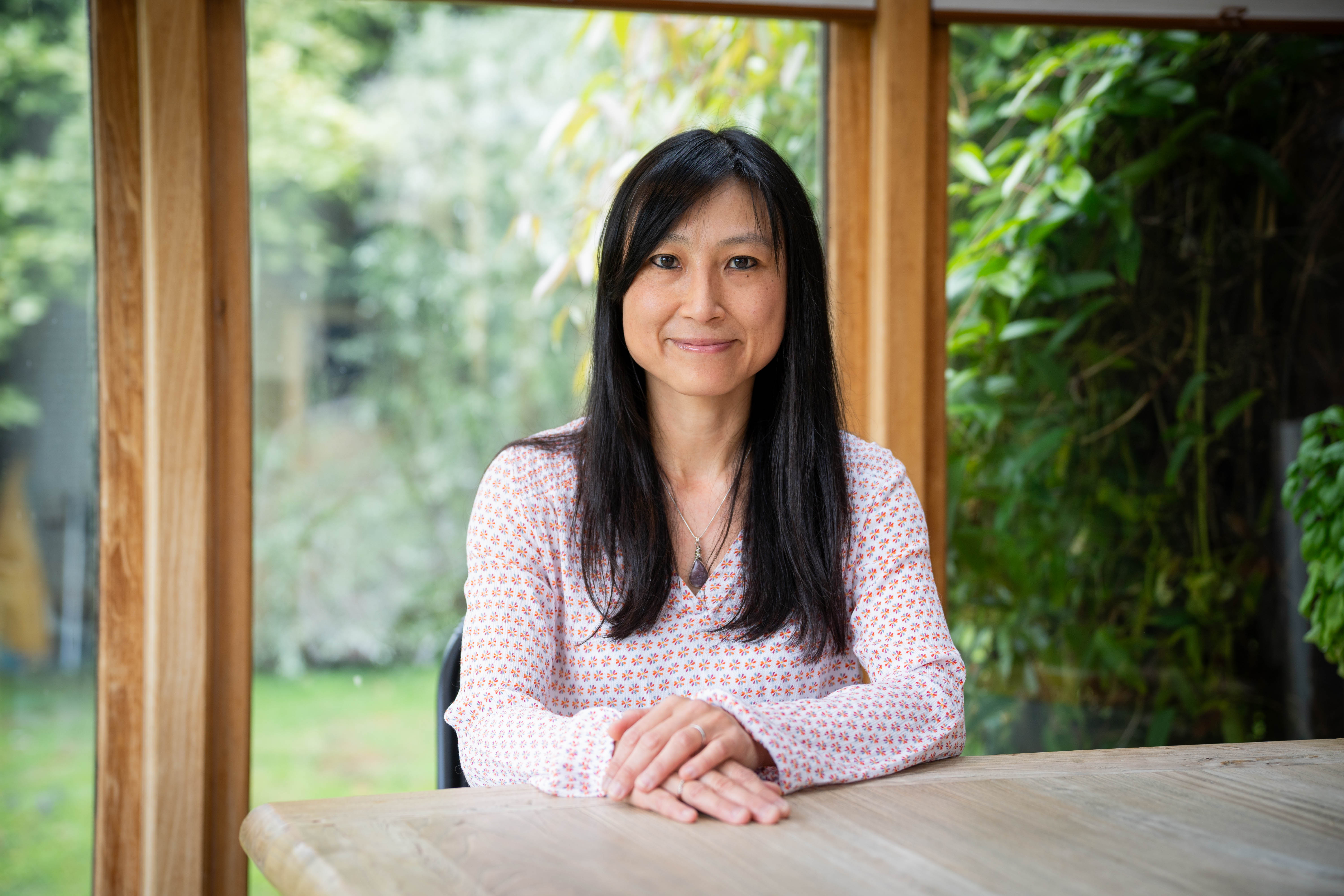 Connie seated at a wooden table in her sunroom, soft daylight through the window frames, garden greenery behind her